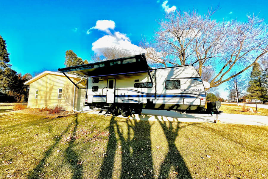 A white Springdale travel trailer with awning extended, parked on a sunny driveway beside a house. Perfect for RV rental.