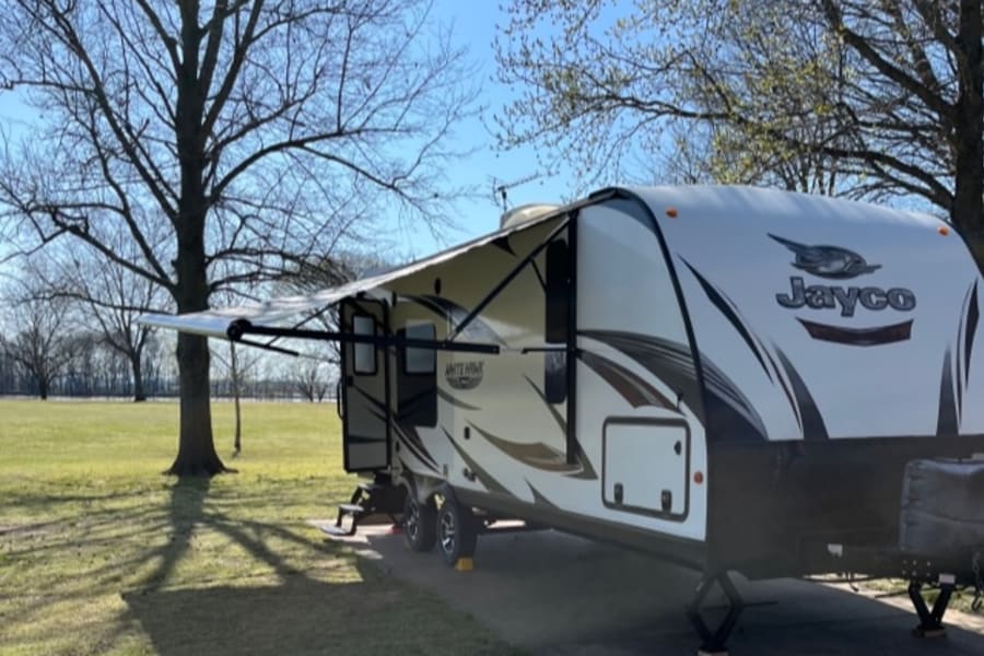 An RV rental, a Jayco travel trailer, is parked at a sunny campsite with its awning extended amidst trees.