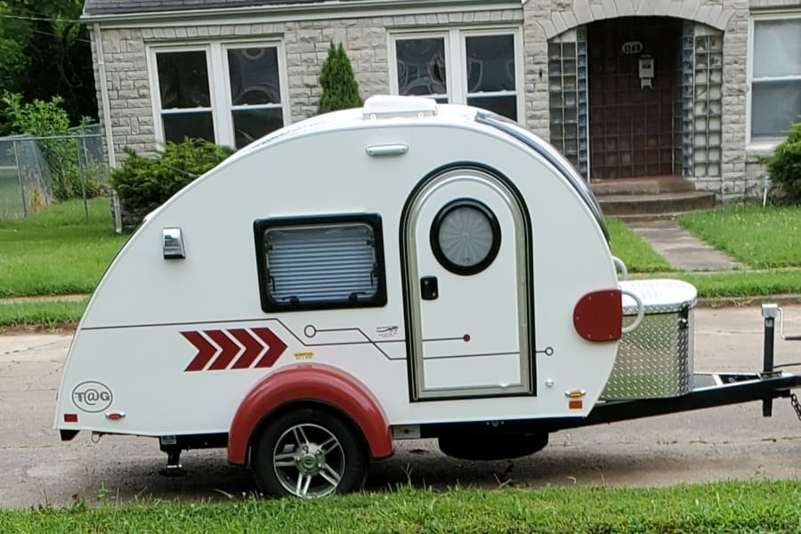 This compact white teardrop camper, perfect for an RV rental, is ready for travel, hitched to an SUV on a sunny street.