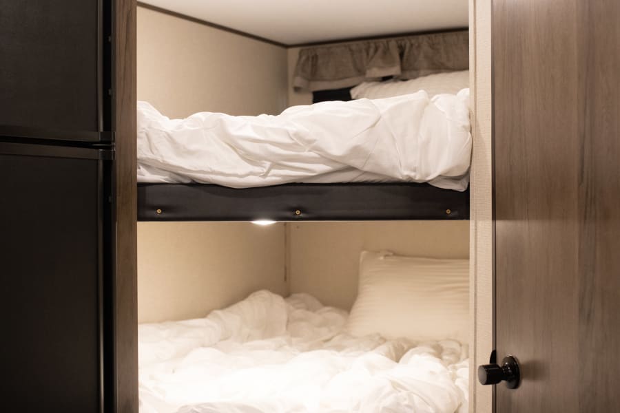 Our RV rental features cozy bunk beds with white bedding, plus a fridge and storage, perfect for your adventure.
