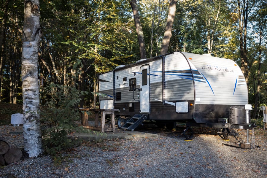 This RV rental, a Prime Time Avenger travel trailer, is parked at a serene, autumn campsite among tall trees.