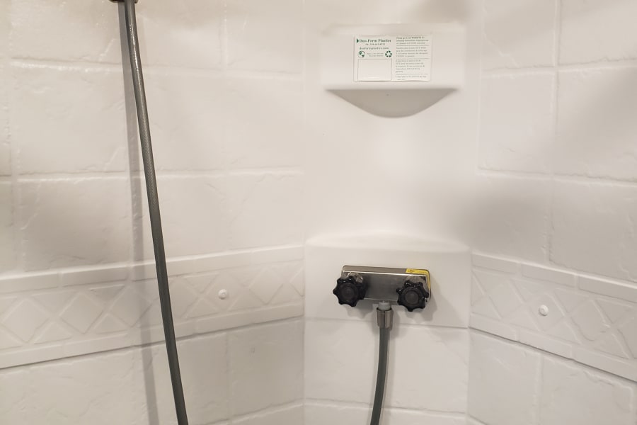 Compact white RV rental shower with handheld showerhead, dual controls, and an integrated shelf for toiletries.