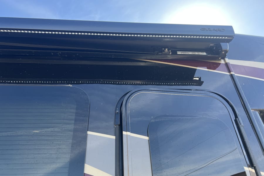 An RV rental's side with a "GIRARD" awning and integrated LED strip lighting. Dark windows with blinds and decorative stripes are also seen.