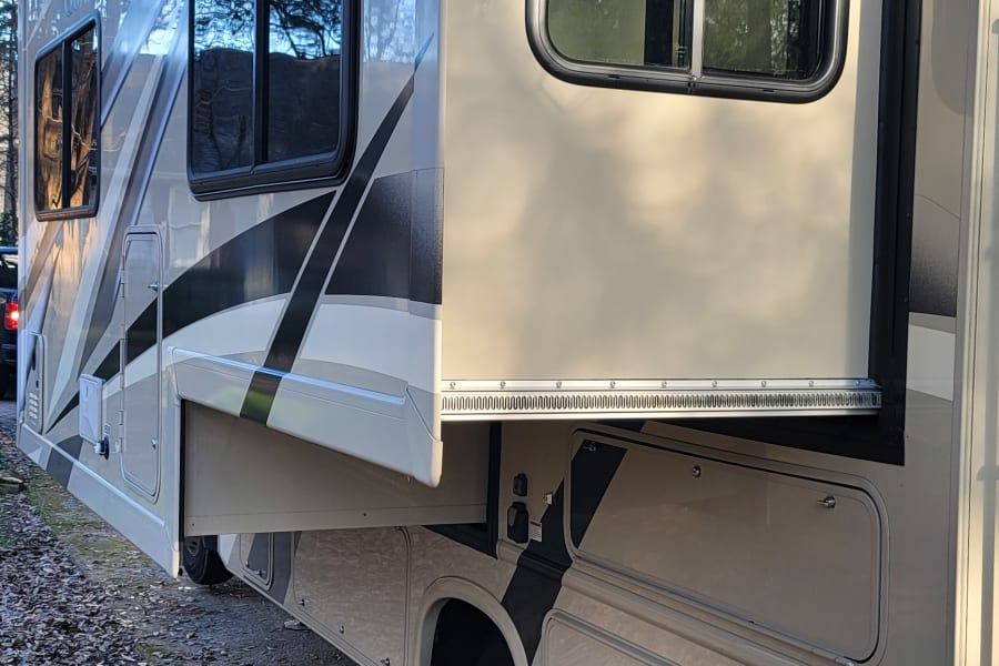 Side view of an RV rental with an extended slide-out and awning, parked on pavement next to autumn leaves.