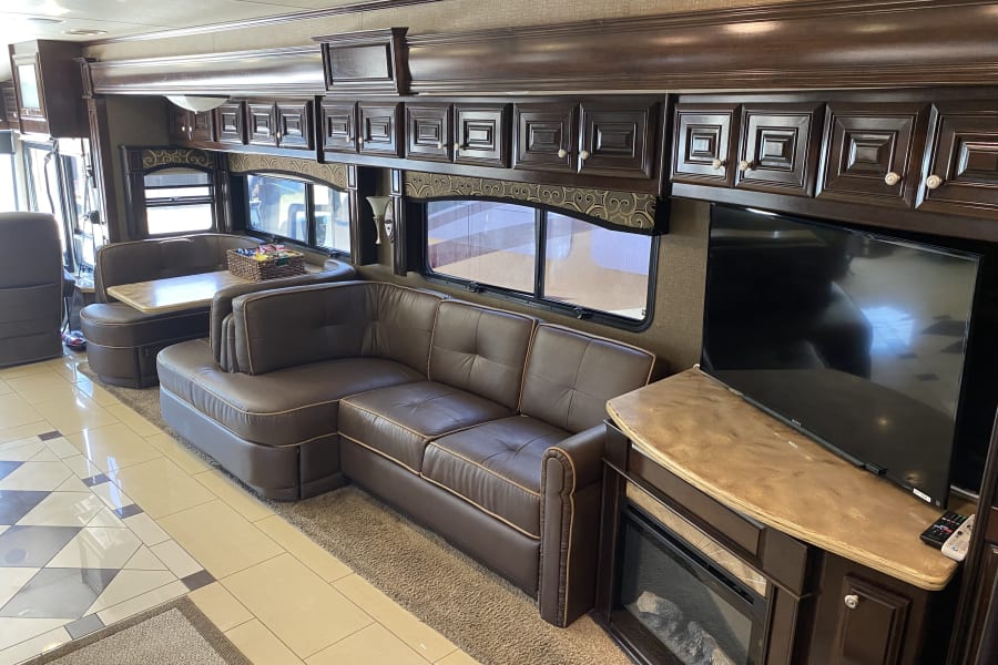 This spacious RV rental interior shows a brown leather sectional, dining booth, TV, and fireplace. Overhead cabinets line the walls.