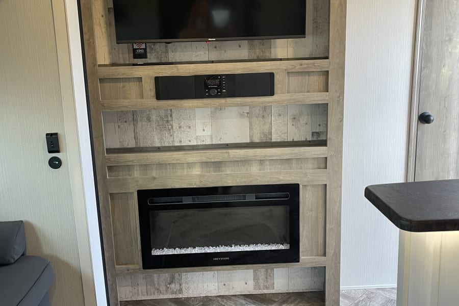 Modern RV rental with an entertainment unit featuring a TV, soundbar, electric fireplace, and wood-look herringbone flooring.