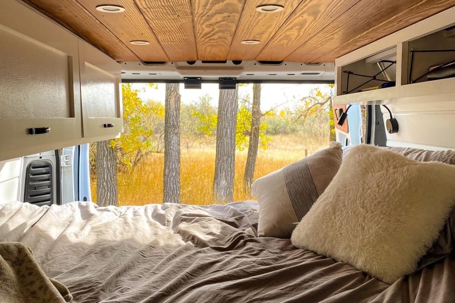 Cozy RV rental interior: a comfortable bed with neutral bedding, wooden ceiling, and a scenic autumn view outside.