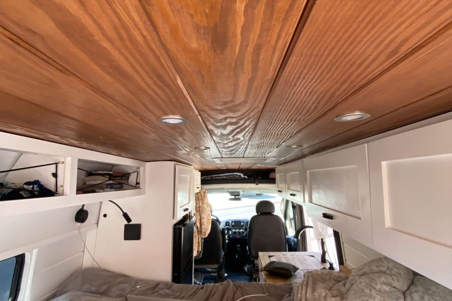 RV rental interior: warm wooden walls, white cabinets, cozy bed with laptop, and a compact kitchen.