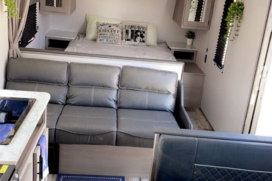 A bright RV rental interior showcases a gray sofa, full kitchen, dinette with marble-look table, and a cozy rear bedroom.