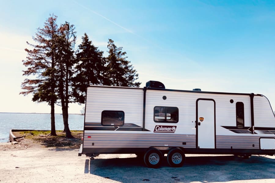 A white Coleman RV rental is parked on a dirt lot by a calm lake with trees under a blue sky.