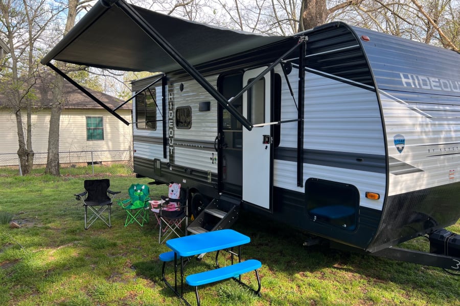 An RV rental, a travel trailer with an extended awning, outdoor chairs, and a blue kids' picnic table on green grass.