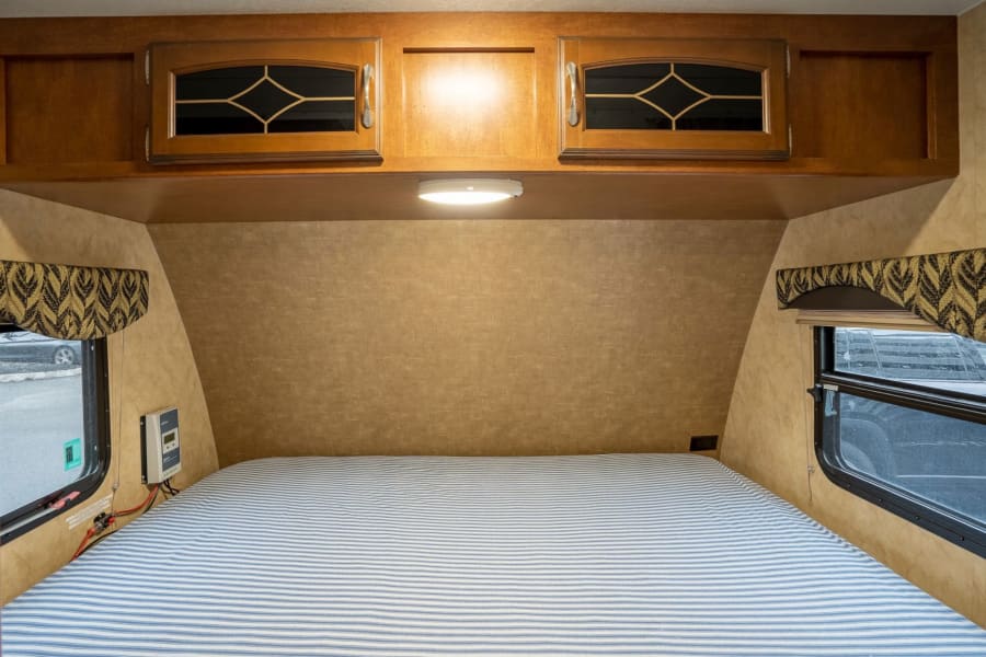 RV rental bedroom: a comfy striped bed, wood cabinets, side windows, and solar charge controller on the wall.
