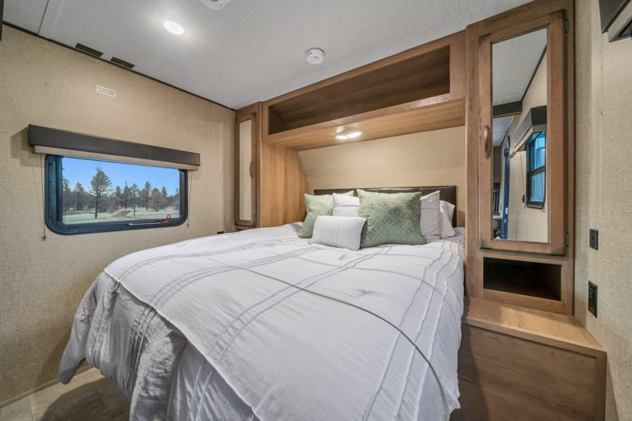 Modern RV rental bedroom: queen bed with green pillows, mirrored wardrobe, and window view of trees. Ideal for travel.