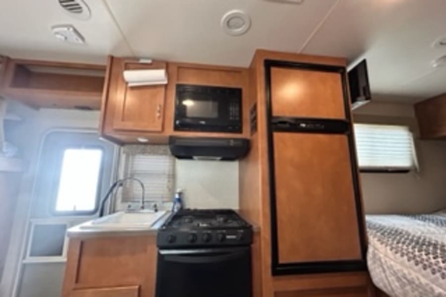 Inside this RV rental, a compact kitchen with wood cabinets, sink, stove, microwave, and fridge offers convenience for travel.