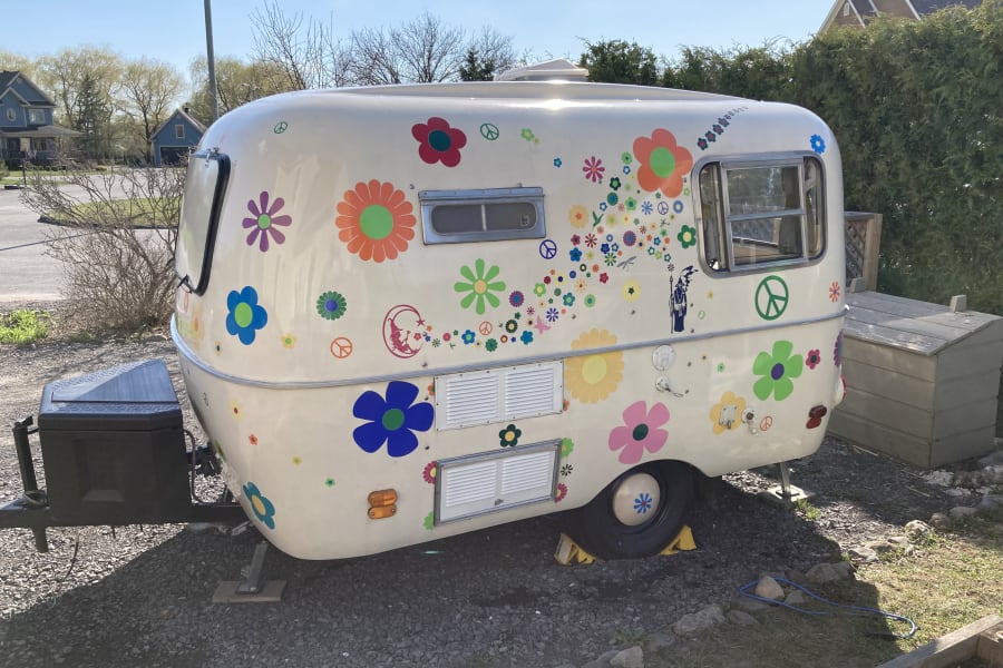 This cheerful, flower-adorned rv rental with peace signs is parked on gravel under a sunny sky, perfect for a fun getaway.