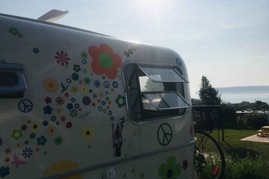 A vintage RV rental, adorned with colorful flower and peace sign decals, set against a sunny, scenic lakeside backdrop.
