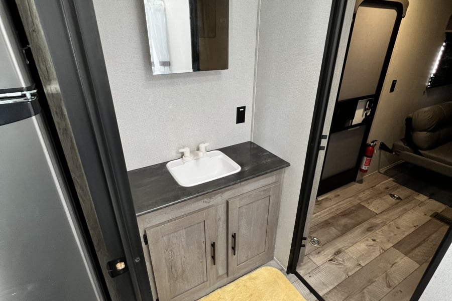 This RV rental features a bright bathroom vanity with a sink, mirror, and yellow mat, leading to the kitchen/living area.