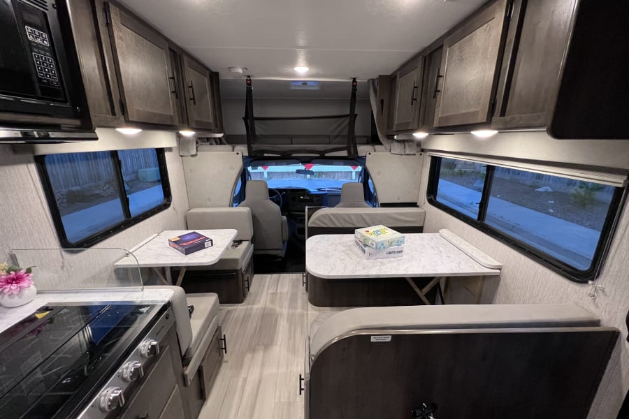 Bright RV rental interior showcasing twin dinettes, a kitchen with microwave/stove, and the front cab area. Games on tables.