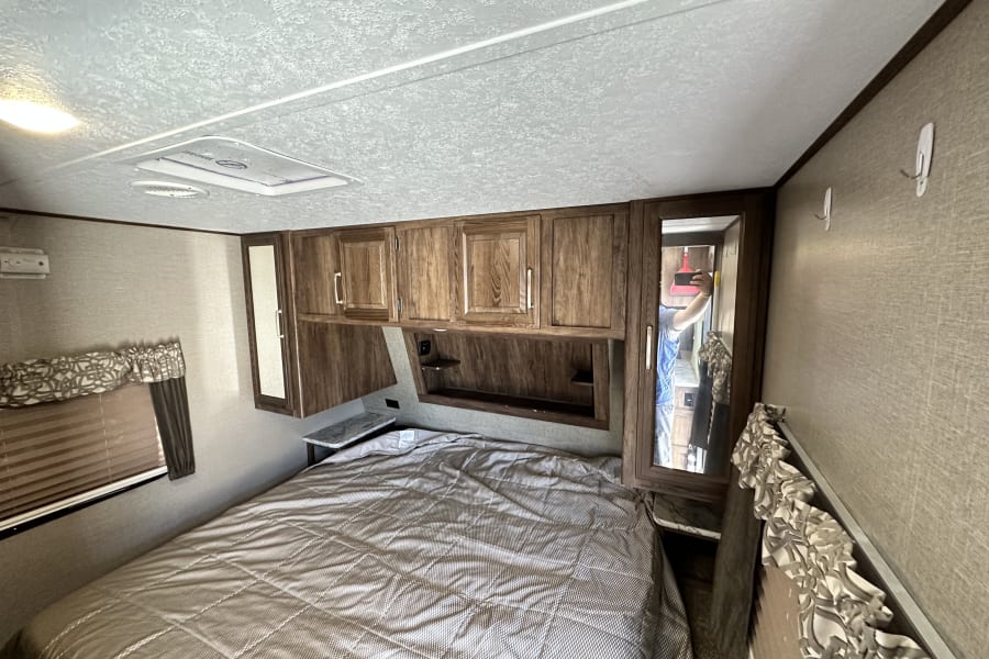 RV rental bedroom features a patterned bed, wooden overhead cabinets, mirrored closet, and window. Great for travel!
