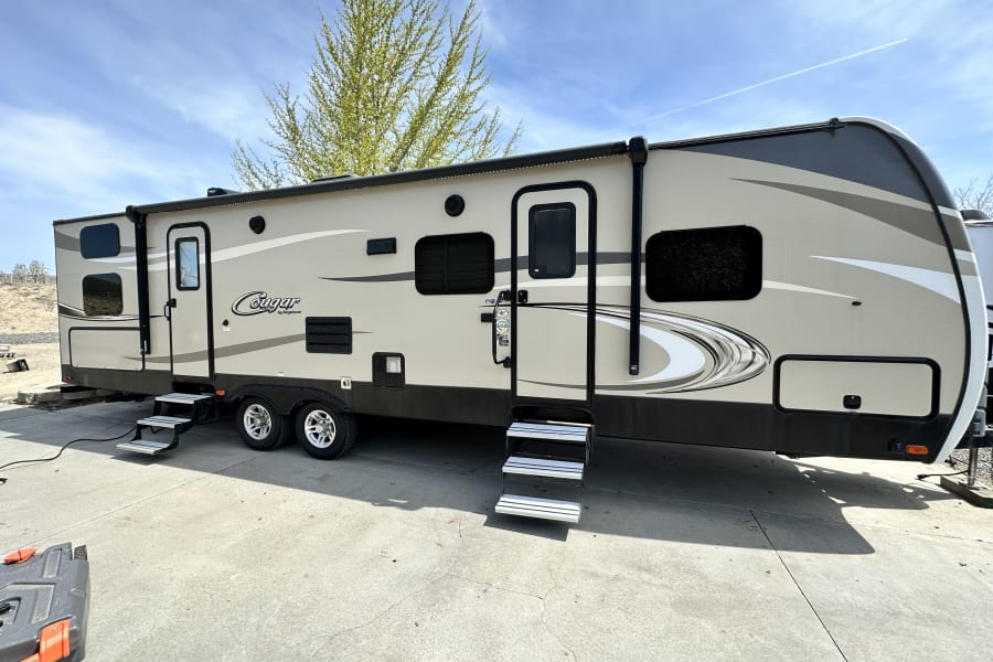 This Keystone Cougar travel trailer, a spacious RV rental, is parked on concrete, ready for outdoor adventures under a blue sky.