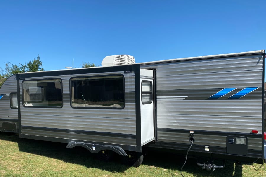 Grey and blue RV rental travel trailer with extended slide-out and roof AC, parked on grass under a clear blue sky.