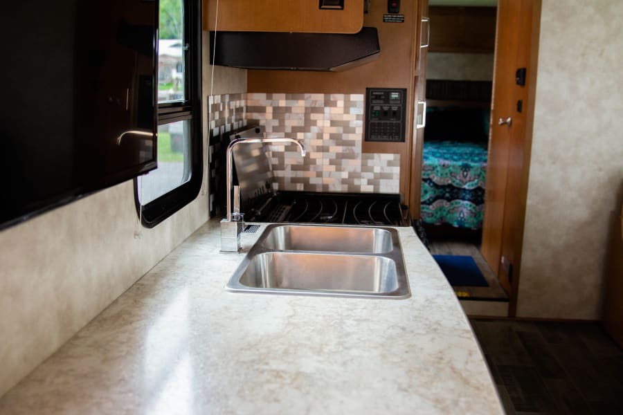 Modern RV rental kitchen with a spacious countertop, double sink, and stove. A comfy bedroom is visible beyond.