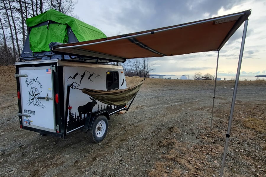 Overland trailer with rooftop tent, hammock, and awning, set up for camping near a body of water&mdash;ideal for an RV rental adventure.