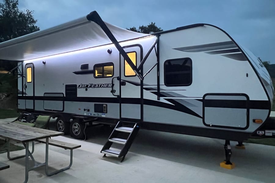 Your perfect RV rental: Jay Feather travel trailer illuminated at dusk, with an extended awning, steps, and picnic table.