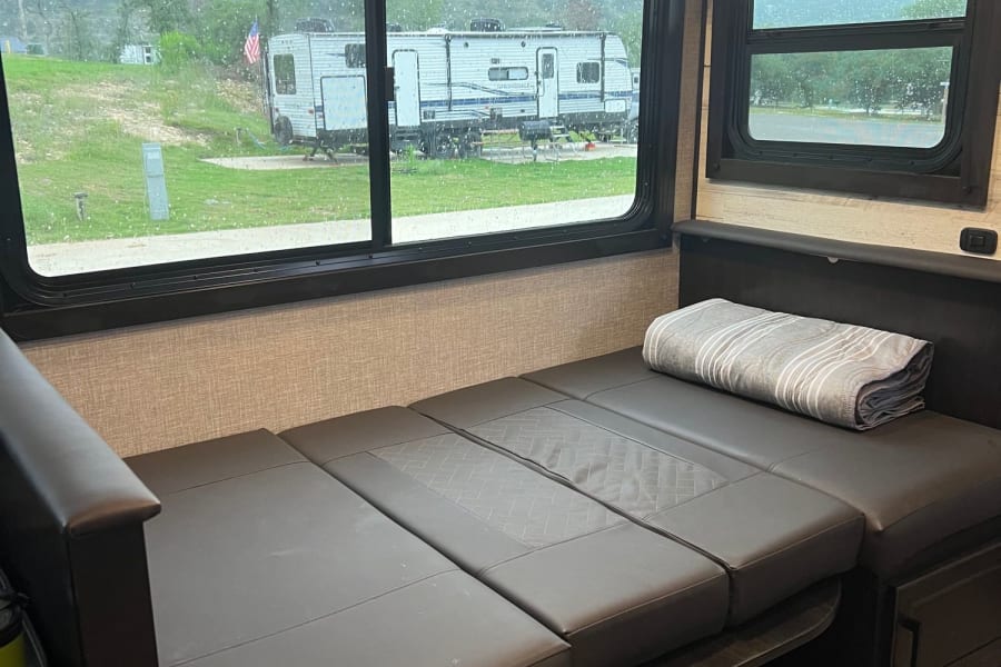 RV rental interior: a dark convertible dinette/sofa overlooks a rainy campground view through large windows.