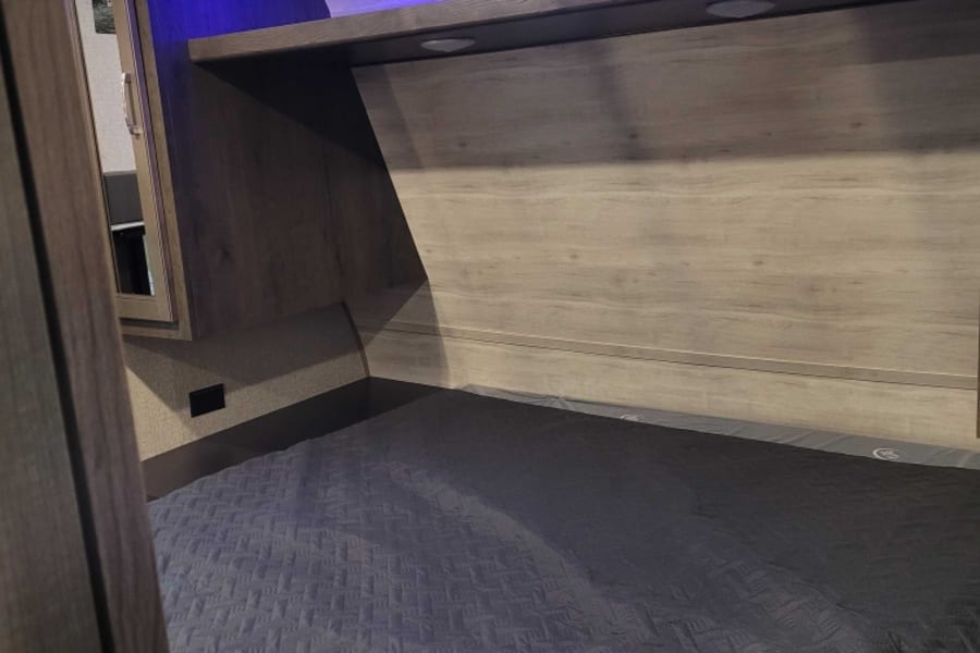 Modern RV rental bedroom with a dark quilted bedspread, overhead shelf, recessed lights, and vibrant blue LED ceiling glow.