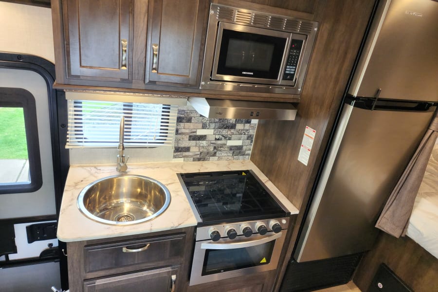 An RV rental kitchenette with a stainless steel microwave, stove/oven, refrigerator, and sink, amidst dark wood cabinets.