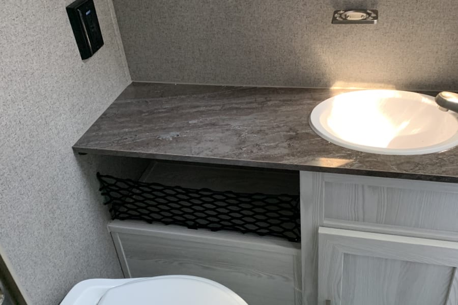 Functional **RV rental** bathroom featuring a toilet, sink, mesh storage, and water jugs. Compact design for travel comfort.
