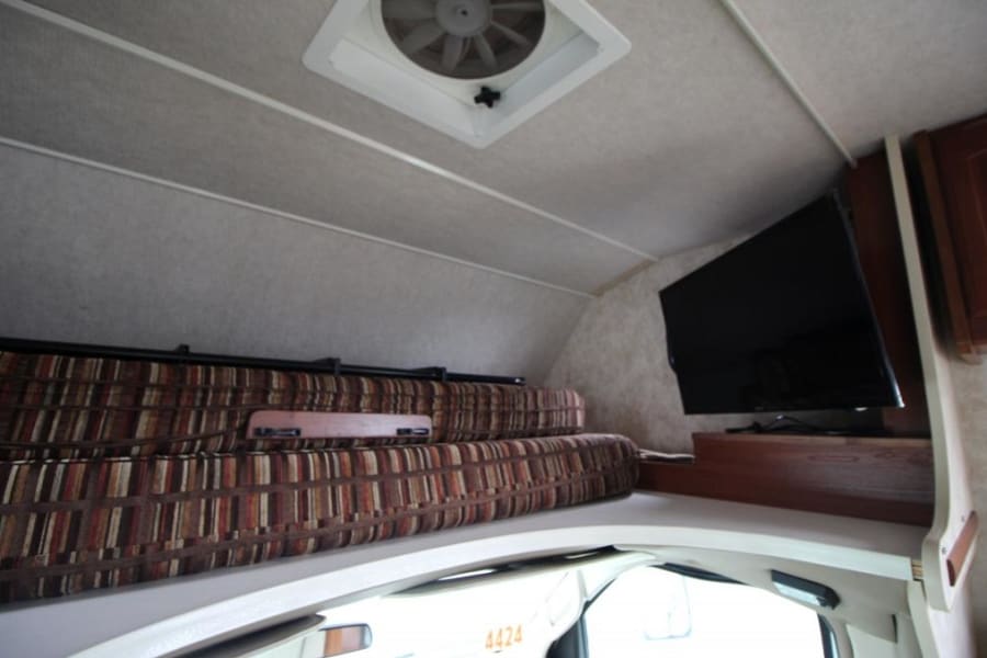 Spacious RV rental cab-over area with comfy striped cushions, wall-mounted TV, and a roof vent for air circulation.