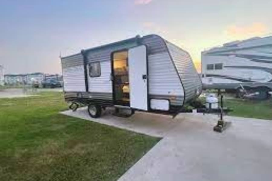 A gray and black RV rental travel trailer with its door open sits on a paved campsite at dusk. Ideal for your next outdoor escape.