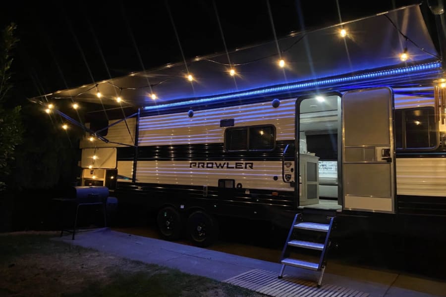 Prowler RV rental at night, illuminated by string lights & blue LEDs under the awning. Steps lead to the open door.