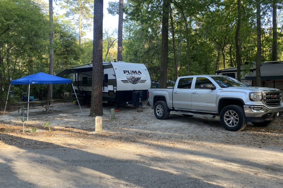 RV rental camping: Silver truck and Puma travel trailer at a sandy campsite with blue canopy, surrounded by tall trees.