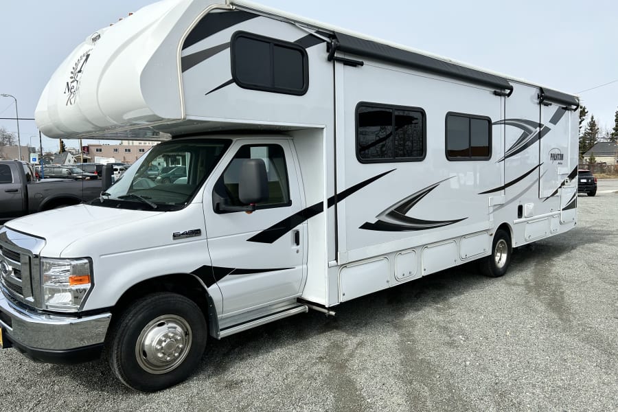 A white Class C rv rental with black/grey graphics, an awning, and multiple storage compartments, parked on gravel.