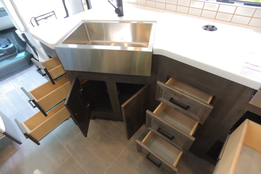 RV rental kitchen: modern stainless steel sink, white counter, dark cabinets with open drawers & doors for storage.