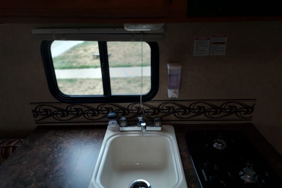 An RV rental's compact kitchen with a sink, two-burner stove, and a window overlooking a campsite, ready for adventure.