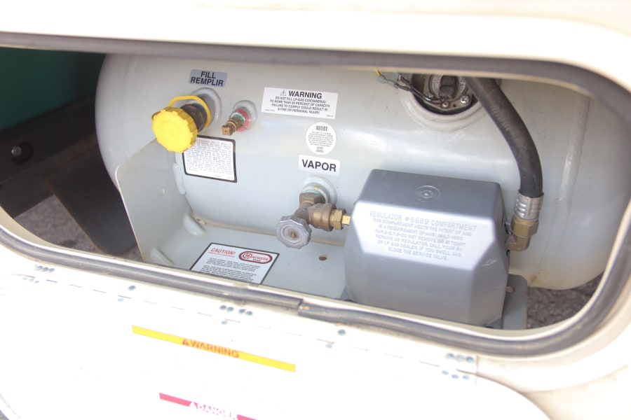 Inside an RV rental's LP gas compartment: a propane tank, fill valve, vapor valve, and safety regulator system.