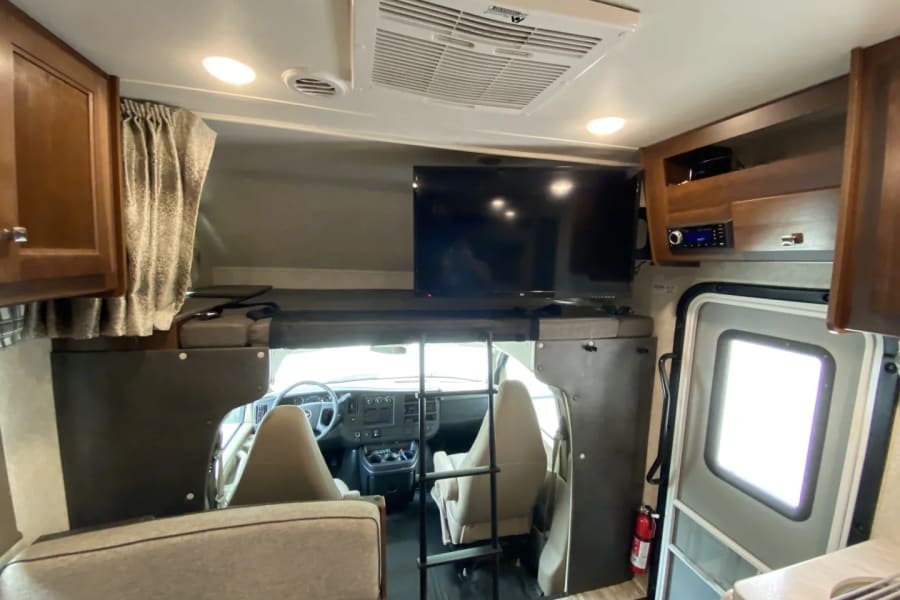 Spacious RV rental interior featuring a cab-over bunk with TV, cockpit, kitchen, and entry door. Ideal for road trips!