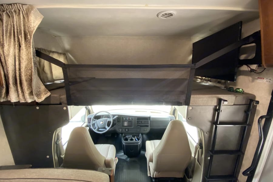 Inside an RV rental: view of the cab-over bunk with safety net, TV, ladder, and the spacious cockpit area.