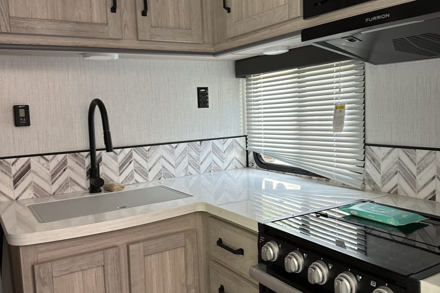 Modern RV rental kitchen featuring light wood cabinets, white counters, sink, stove, and microwave.