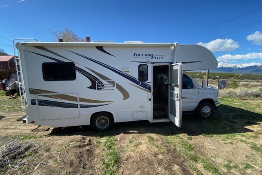 Spacious white Class C motorhome (Freedom Elite) RV rental parked on grass with scenic mountains under blue sky.