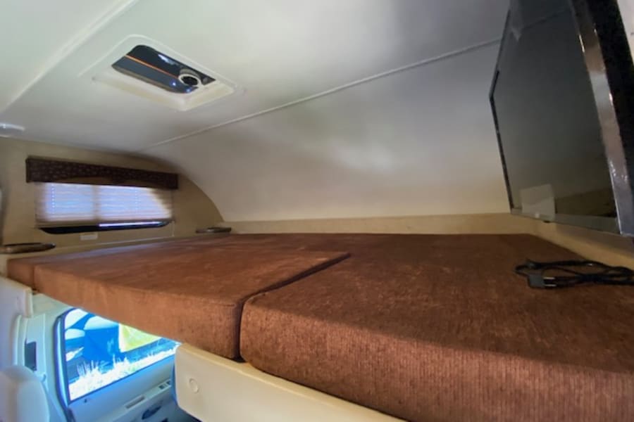 An RV rental's cab-over bunk space, featuring a brown cushioned bed, side window, ceiling vent, and mounted TV.
