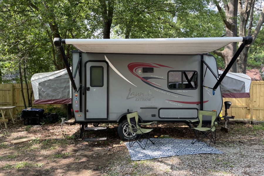 An RV rental: grey travel trailer with pop-outs, extended awning, outdoor chairs, and a patterned rug in a wooded area.