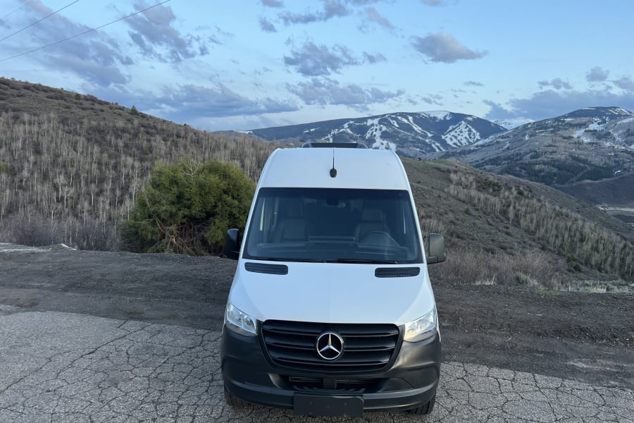 White Mercedes-Benz Sprinter RV rental van on a cracked road, with snow-patched mountains in the scenic background.