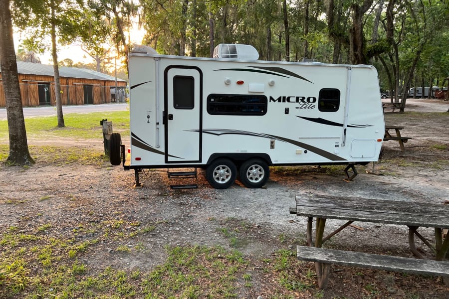 RV rental: A white travel trailer is parked on a shaded, tree-lined campsite with a wooden picnic table.