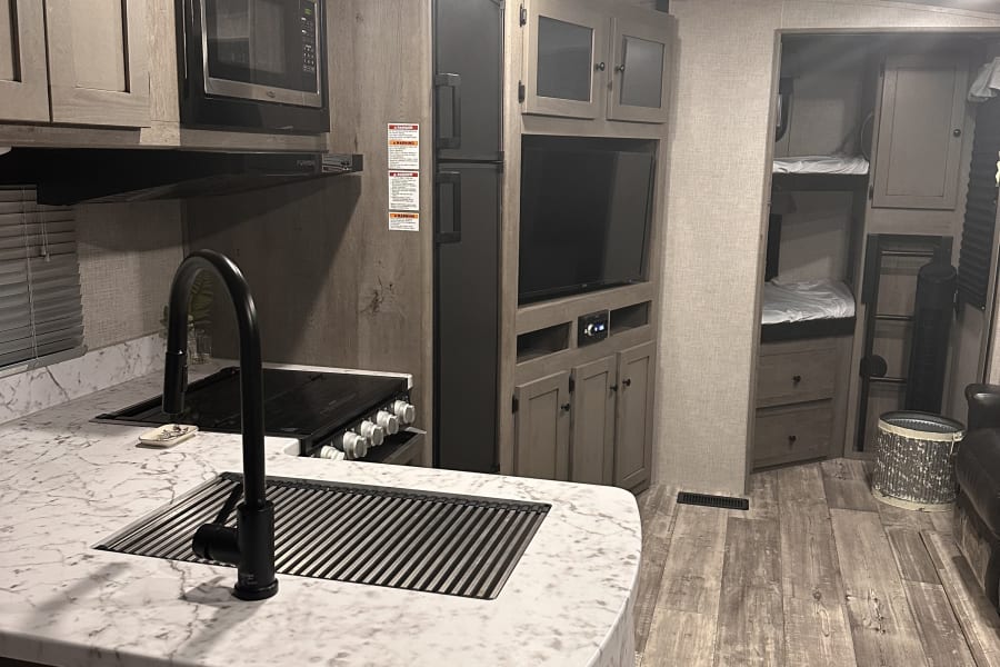 Modern RV rental interior showcases a kitchen with white counters, a living area, and bunk beds for family comfort.