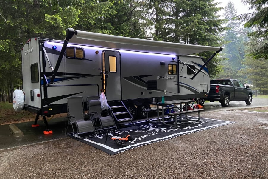 An RV rental parked at a wet campsite with a truck, extended awning, and LED lights, ready for forest camping.